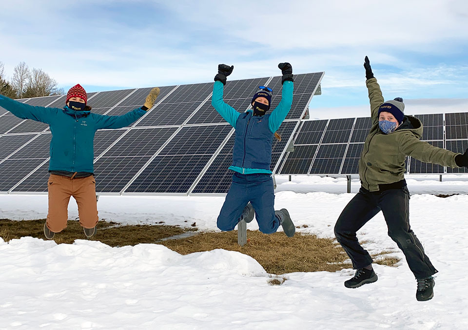Three women are jumping toward the air with arms splayed, in front of large solar panels.